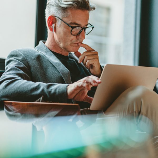 Man wearing glasses and earpods considering something on a tablet computer while sitting in a chair with legs crossed